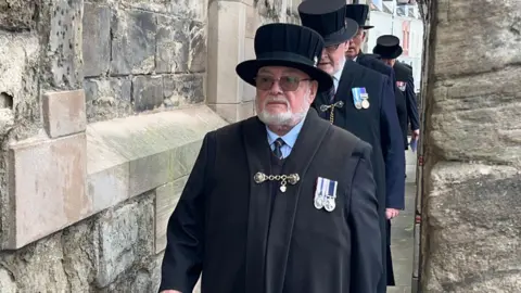 A man wearing a black suit and top hat walks through a brick alleyway.