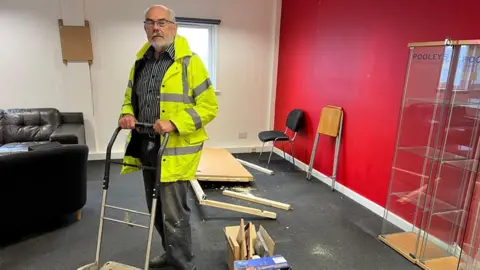 Glyn Matthews A man in glasses and a high-visibility yellow jacket grips a metal sack trolley in an emptied office. Dismantled flat-pack furniture and a cardboard box lie on the dark carpet around him. A bright red wall and empty glass display cabinets stand behind him.