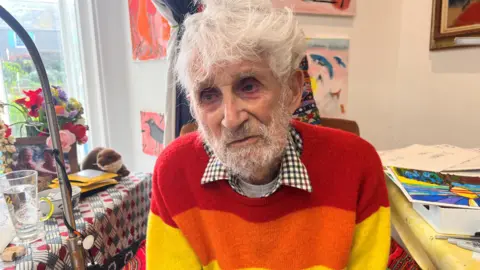 Philip Sutton, 96, is looking at the camera, has white hair and a grey beards. He is wear a checked shirt with a bright coloured jumper over the top with is art studio in the background.