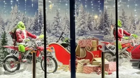 MG Accident Repair Centre The festive window scene shows two Grinches on red motorbikes either side of a sleigh with fake snow on the ground and presents near to it. The end of a red car can be seen on the right.