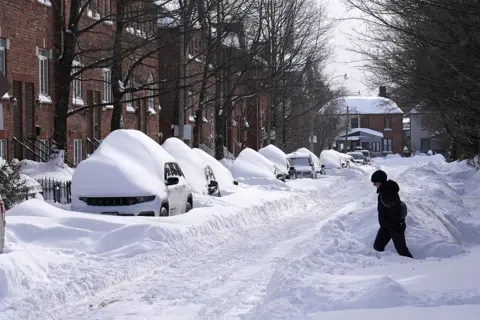Anadolu via Getty Images Mieszkaniec Toronto w Kanadzie przechodzi przez zaspy głębokiego śniegu. Na ulicy stoją samochody zasypane śniegiem.