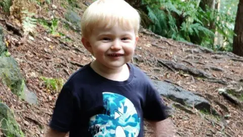 Edward smiling at the camera with blonde hair and wearing a navy blue t-shirt with a light blue pattern. He is stood in woodlands. 