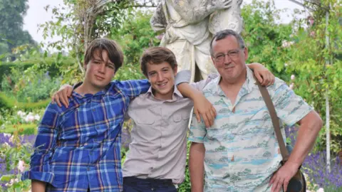 Family photo A man with grey hair and a white patterned shirt standing next to two young men with brown hair and shirts in front of a stone statue and bushes