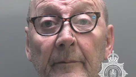 NWP James Hughes police mugshot. He has stubble and is wearing spectacles