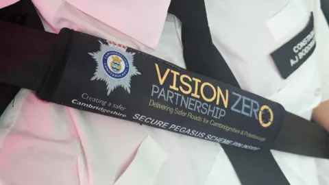 Cambridgeshire Police Image shows a police officer wearing the black seatbelt cover, which says on it in blue and yellow writing: "VISION ZERO PARTNERSHIP, Delivering Safer Roads for Cambridgeshire & Peterborough. SECURE PEGASUS SCHEME PIN INSIDE". There is the Cambridgeshire Police logo on it.