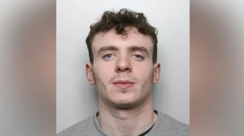 Wiltshire Police A mugshot of a 20-year-old man. He has dark brown hair which is short on the sides and curly on the top, bushy eyebrows and minimal facial hair. He appears to be smirking slightly. He is wearing a grey sweatshirt and is standing in front of a pale, plain background.