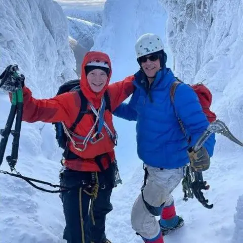 Kym McGillicuddy Two people smile on a snowy mountain with climbing gear, the one on the left wears a red jacket and the one on the right wears a blue jacket