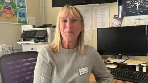 BBC Melanie Dawson, with long blonde hair, is sitting facing the camera with her back to the desk. It has a computer screen, which is turned off, and keyboard. She is sitting on a purple chair.