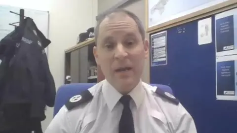 A police officer faces the screen. He wears a shirt, tie and epaulettes. An office is visible in the background. A police uniform coat hangs on a stand.  