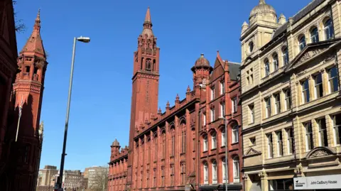 A red brick and terracotta building with a tower, on a sunny street.