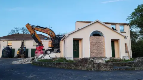 The remains of Clonduff parochial house. The front section of the two-storey building is boarded up and a yellow mechanical digger is behind the partial structure. More heavy plant equipment is parked near the rear of the site.