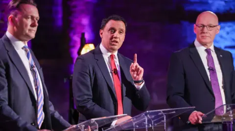 PA Media Three men in suits stand at lecterns during a debate; the central speaker in a red tie raises a finger while speaking, flanked by two others, against a purple-lit brick backdrop.