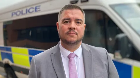 Det Ch Insp Stuart Hodgson wears a grey blazer, white shirt and pink tie, with short dark/greying hair and a stubble moustache and beard, and looks into the camera. A police van is parked behind him.