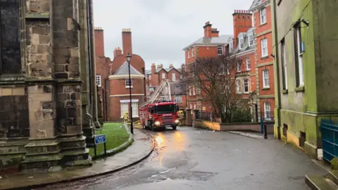A red fire engine on a narrow street next to red brick buildings. There are white and blue police cordons nearby