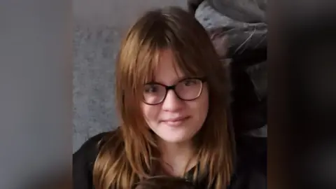 Natalie-Anne Rowlett Felicity-Jo before illness - she smiles and has brown hair with a fringe and wears black framed glasses.