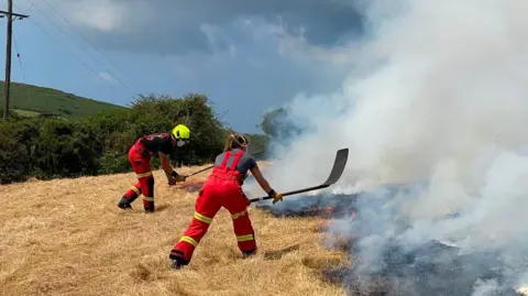 Cornwall Fire and Rescue Service Two firefighters in red uniform lean over a burnt out patch of grassland to put out a fire in broad daylight, as thick black smoke rises into the air.