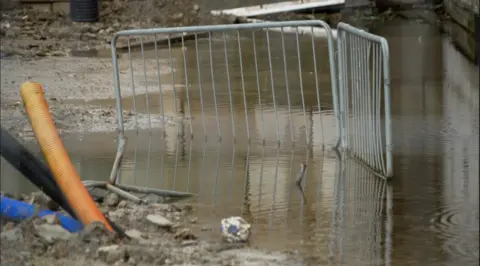 A puddle of water next to the breeze block. Close to the water are pipes coming from out of the ground. A piece of portable metal fencing is part submerged in the puddle.