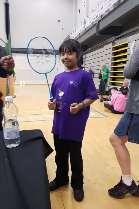 A young girl holding a blue badminton racket smiles into the camera. She is wearing a purple t-shirt and black trousers with black trainers. In the background there is a busy sports hall. 