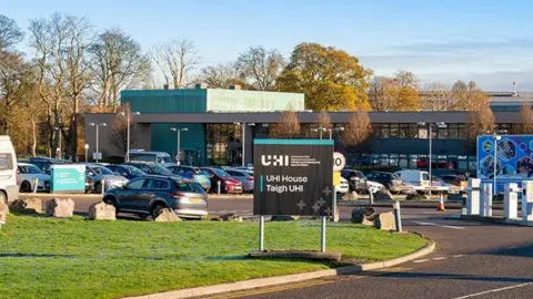 UHI House is a glass-fronted building with a green box-shaped feature. There is a busy car park in front of it an sign that reads "UHI" and "UHI House" in English and Gaelic.