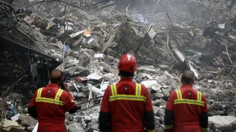 Getty Images Three rescue workers stand facing a pile of rubble. 