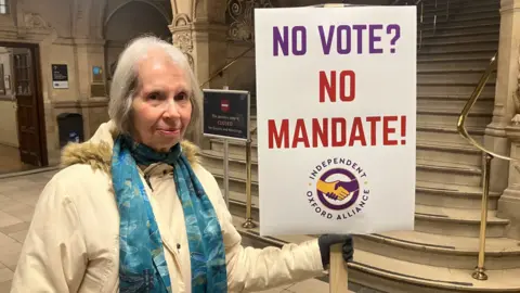 Anne Gwinett from the Independent Oxford Alliance stands in the lobby of the town hall in Oxford. She is carrying a sign that says "No Vote? No Mandate!"