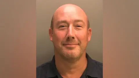 A mug shot of a man looking into the camera. He is bald with light stubble. He is wearing a dark blue collared shirt. 