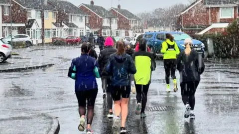 Runclub Women in running and outgear gear are running through sleet in a residential street. There are houses on both side of the road. The women are in a group in the middle of the road.