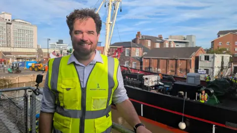 A man with curly short brown hair is wearing a yellow hi-vis jacket and a white shirt and smiling into the camera as he squints a bit in the sun. He is standing on an elevated balcony, with a trawler behind him in a built-up city area.