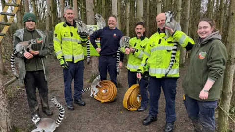 A group of men and a woman stand in a woodland. Three of the men are wearing hi-vis jackets used by the fire serice and are holding lemurs on their arms or on their shoulders. Another lemur sits on the floor, with one lemur sitting on an orange fire hose in the background.