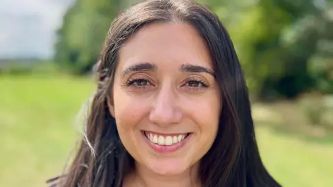 Shanika Mahendran, standing outside, smiling at the camera, with long dark hair. Trees and grass is behind her, but blurred. 