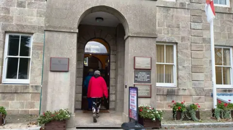 Woman in a redcoat entering the island hall in Alderney. The large stone building has an arched entrance.