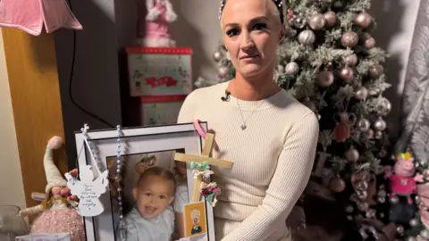 Natasha Broadley, facing camera, in front of a Christmas tree, holding a large framed photograph of her daughter Louisiana Brooke Dolan