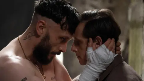 A man with a beard and bandages on his hands holds the face of a man wearing a suit.