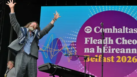 Pacemaker Press A man is standing onstage with his arms up. He has long hair and a beard and wears glasses. He's wearing a grey suit. 