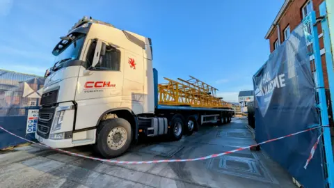 Simon Pattern A lorry carrying parts of the crane. The low-loader articulated lorry has a white cab. It is carrying the bright yellow parts of the crane. 
