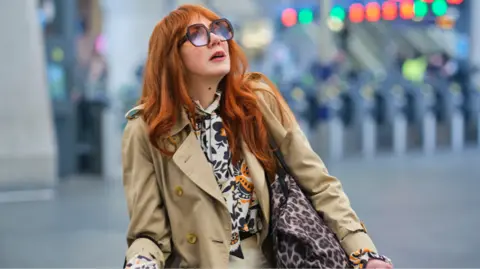Govia Thameslink Railway Diane Morgan wearing big glasses looking up with railway station barriers behind her