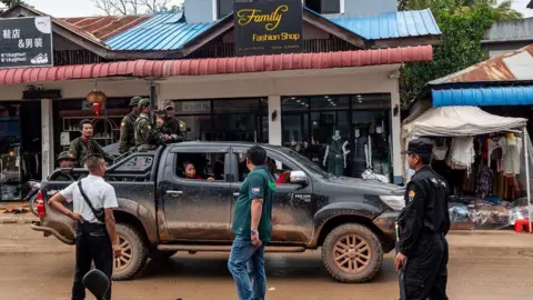 Jonathan Head/ BBC A vehicle inside the city. Armed men who work for the local warlord in fatigues stand on the back of a black pickup truck driving down a road with shops on it, one called Family Fashion Shop