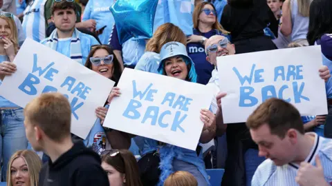 Coventry City fans dressed in Sky Blue in the stands of the Coventry Building Society Arena. the image centres on three fans holding up We Are Back signs. The woman in the centre has sky blue dyed hair and a blue bucket hat. On her right is a man with heart-shaped sky blue glasses and on her left a woman in the same glasses with long dark hair.