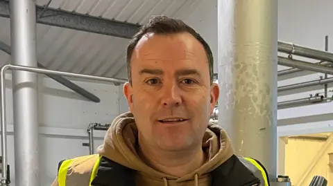 James Ramm is wearing a hi-vis jacket and a light brown hoodie. He is standing in a brewery, surrounded by stainless steel brewing vessels, and is holding a bottle of beer.