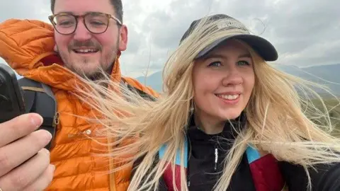 Rachael Dunfell A woman wearing a baseball cap smiles on a hike, standing beside a man wearing an orange jacket.