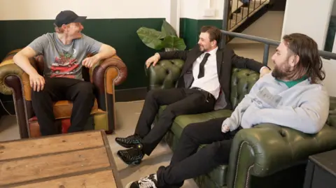 Three young men are sitting on leather furniture in a common room talking to each other. The man on the right is wearing a grey shirt, black trousers and a black cap. He is smiling at the second man, who has slicked-back brown hair and is sitting on a green sofa and wearing a black suit with blue trainers. On the right, the third man has long brown hair and a full beard and is wearing a grey shirt and black trousers. 