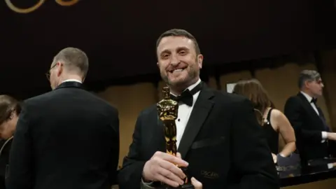 Mike Hill, wearing a black tuxedo, smiles as he hold an Oscar backstage at the Oscars ceremony