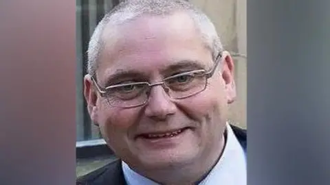 Lancashire Police David Daintree smiles into the camera. He has short grey hair and glasses and is wearing a shirt and tie and suit. 