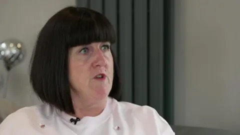 BBC News Maura Jackson, a middle-aged woman, sits speaking to someone off camera. She has a dark bob and wears a white t-shirt. 