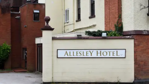 The words Allesley Hotel are in large letters on a light-coloured wall facing the camera. Two doors on the left of the photo at ground level are at an angle to the camera, which is outside.