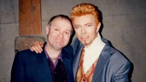 Two men are looking into a camera and smiling for a head and shoulders shot. The man on the left has short brown hair and is wearing a blue blazer and purple shirt unbuttoned a bit. On the right is singer David Bowie, who has orange hair spiked up, short stubble on hic chin, is wearing a blue jacket and patterned red and yellow waistcoat with a white top under it. He has his arm around the man next to him.