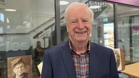 Seamus McKee smiles at the camera, he has short white hair and is wearing a dark blue suit jacket over a checked shirt. He is standing in front of a glass partition in Falls Road Library.