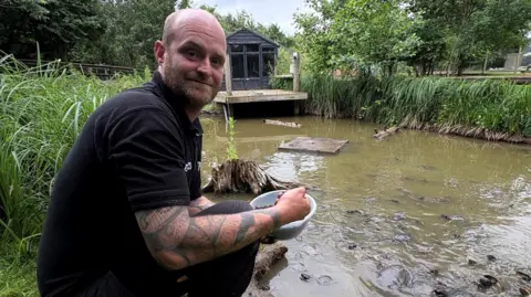 Kevin Shoesmith/BBC A man wearing a black T-shirt squats next to a pond filled with terrapins. He has his hand in a plastic bowl containing food pellets.