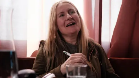 Viv Gee sitting at a table with a pen in her hand. She has long, light brown hair and is wearing a khaki hoodie.
