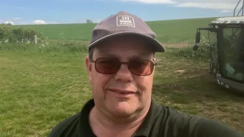 A selfie of a male farmer who is standing in a field. His tractor is next to him. He is wearing a blue hat and a green top. There is a green field behind him. 